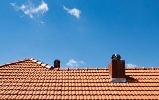 A red tiled roof with two attic vents and a chimney stands against a clear blue sky with a few small clouds, prompting the question: How Long Should a Roof Last?.