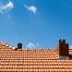 A red tiled roof with two attic vents and a chimney stands against a clear blue sky with a few small clouds, prompting the question: How Long Should a Roof Last?.