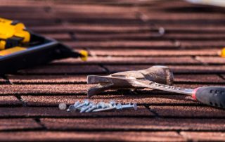 A close-up of a hammer and nails on a shingle roof, with a yellow nail gun in the background.