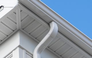Close-up of the corner of a house roof showing white soffit, fascia, and a downspout—an example often seen when exploring different types of gutters—against a clear blue sky.