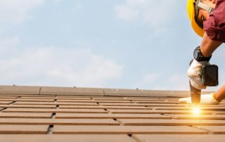 Construction worker in safety gear using a power tool to install shingles on a roof during a roof replacement under a clear sky.