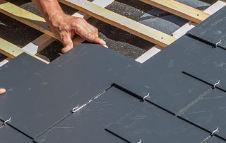 A person installs rectangular slate tiles on a wooden roof structure during a roof replacement, using metal hooks to secure each tile in place.