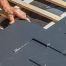 A person installs rectangular slate tiles on a wooden roof structure during a roof replacement, using metal hooks to secure each tile in place.
