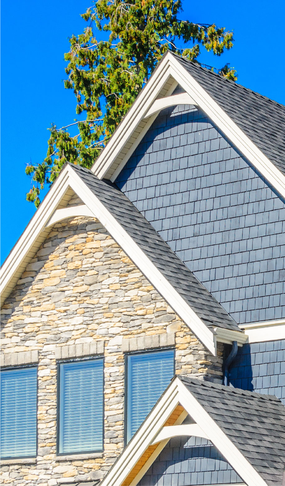 Close-up of a house exterior featuring grey shingle siding, stone veneer, and white trim under a clear blue sky with a tree partially visible—ideal for documenting insurance claims.