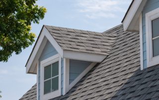 Close-up view of a house roof with gray asphalt shingles after roof replacement, featuring two dormer windows and a partial view of a leafy green tree.