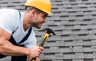 A construction worker wearing a yellow hard hat and overalls uses a hammer while working on a rooftop with asphalt shingles during a roof replacement.