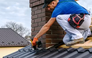 A worker in a blue shirt installs metal roofing sheets around a brick chimney on a house roof undergoing roof replacement.