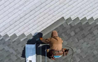 A worker in a brown hoodie installs shingles on a roof during a roof replacement, using tools and materials placed nearby, viewed from above.