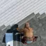 A worker in a brown hoodie installs shingles on a roof during a roof replacement, using tools and materials placed nearby, viewed from above.