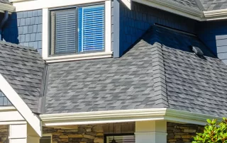 A modern two-story house featuring stone and blue siding, multiple gabled roofs with dark shingles that may answer your roofing questions, white trim, and several windows with blue shutters.