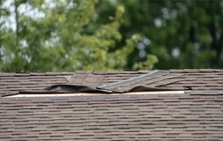 Close-up of a roof with damaged and lifted asphalt shingles, exposing the underlayment beneath—evidence of storm-related damage with trees blurred in the background.