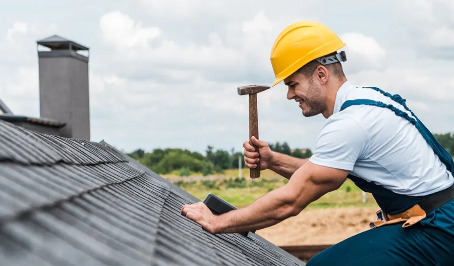 A construction worker in a yellow hard hat and overalls, representing a Roofing Contractor in Winchester, uses a hammer to work on a shingled roof outdoors.